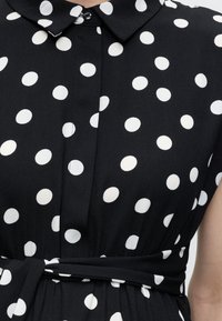 Must valge täpilise mustriga kleit, kaelusega, nööpidega ja vööna seotud kangariba vöökohal.