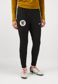 Sorte treningsbukser med et løveemblem på låret, Nike-logo og gule sideaksenter. Laget av glatt, lett materiale.