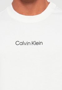 Nærbilde av en person som har på seg en hvit skjorte med "Calvin Klein" trykket i svart sentrert på brystet.