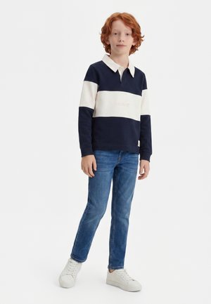 Enfant roux debout, portant un polo à manches longues bleu marine et blanc, un jean bleu et des baskets blanches sur un fond uni.