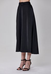 Jupe noire en forme A en satin, dotée d'une large ceinture à la taille et d'une texture lisse. Associée à des sandales à talons hauts noires et à brides.