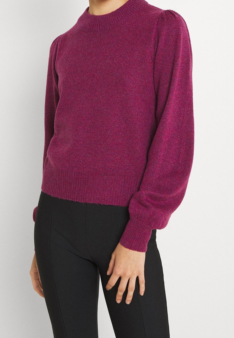 Femme portant un pull en maille violet à manches longues côtelées avec des épaules bouffantes, associé à un pantalon noir ajusté.