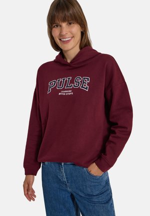 Femme souriante portant un sweat à capuche bordeaux avec le texte "PULSE learning never stops" et un jean bleu, debout avec une main dans la poche.