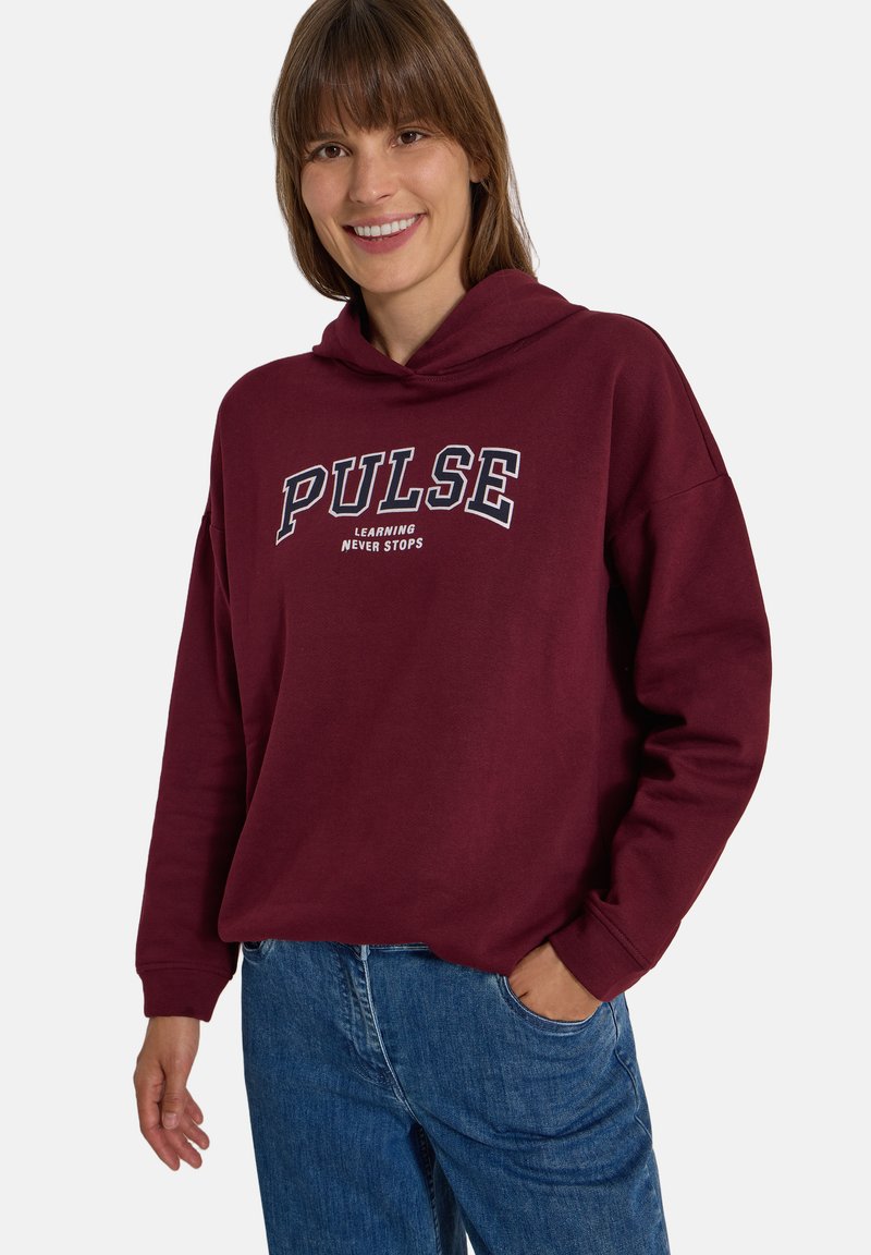 Femme souriante portant un sweat à capuche bordeaux avec le texte "PULSE learning never stops" et un jean bleu, debout avec une main dans la poche.