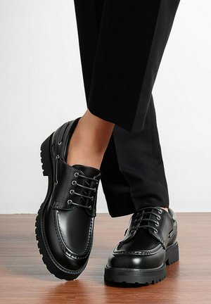 Chaussures noires en cuir à lacets avec semelles épaisses portées avec un pantalon noir, posées sur un sol en bois.