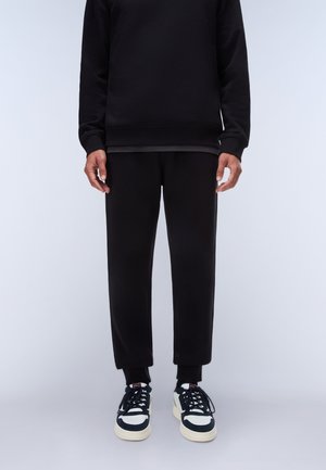 Persona che indossa pantaloni neri, felpa nera e sneakers bianche e nere, in piedi davanti a uno sfondo chiaro uniforme.