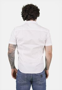 Camicia bianca a maniche corte con colletto classico, caratterizzata da una texture liscia e dettagli di pieghe sul retro. Abbinata a jeans di denim blu.