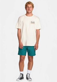 Crème katoenen T-shirt met een bergafbeelding op de zak en turquoise shorts. Model draagt zwart-witte high-top sneakers met witte sokken.