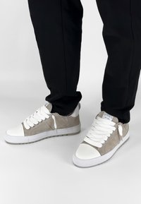 Beige suède sneakers met een witte leren neuskap, dikke witte veters en een gestructureerde zool. Label zichtbaar op de hiel; gestyled met zwarte broek.