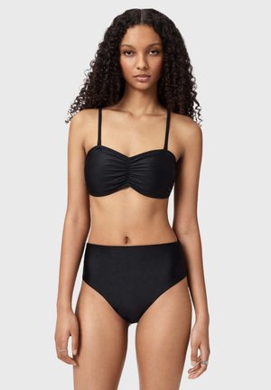 Ung kvinde med langt krøllet hår iført en sort rynket bandeau-bikini top og bikinitrusser med høj talje, stående mod en grå baggrund.