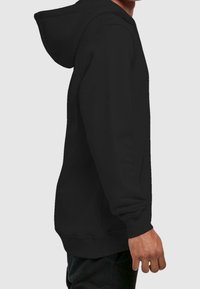 Vista lateral de una persona que lleva una sudadera negra sencilla con mangas largas y capucha, contra un fondo gris claro.