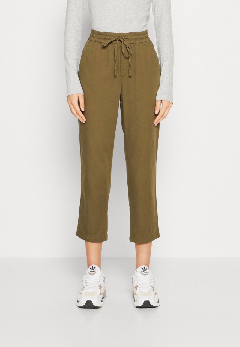 Pantalons en toile khaki, coupés au-dessus de la cheville, avec une taille élastique et un cordon de serrage, présentant une coupe décontractée. Portés avec des baskets blanches et crème.