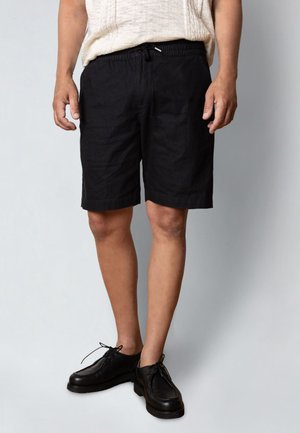 Mann iført svarte shorts med snor, svarte snøresko og en lys beige teksturert kortermet skjorte, stående mot en enkel bakgrunn.