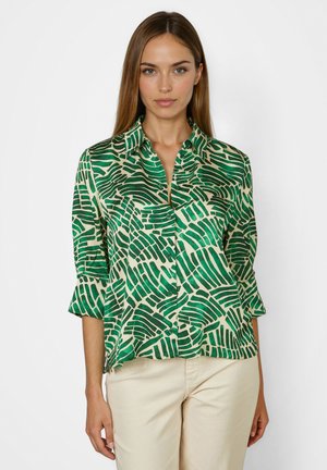 Woman wearing a green and white patterned button-up shirt with three-quarter sleeves and beige pants, standing against a plain background.