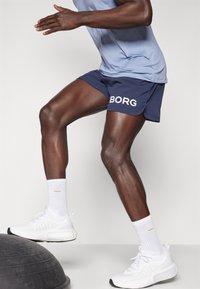 Calças de desporto em azul-marinho com o logótipo branco "BORG", combinadas com ténis brancos e meias até ao tornozelo. Modelo está na posição de corrida sobre uma bola de estabilidade.