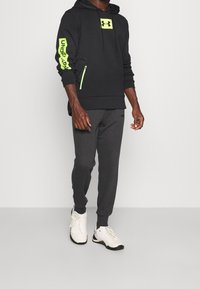 Felpa nera con dettagli verde fluo, dotata di tasca con zip e logo. Abbinata a pantaloni sportivi grigi scuri e scarpe da ginnastica bianche.