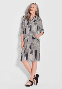 Graues Kleid mit geometrischen schwarz-weißen Mustern, mit Kragen, dreiviertelÄrmel, Taschendetails, kombiniert mit schwarzen Keilsandalen.