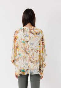 Blusa floreale trasparente con grandi motivi di fiori beige e pastello, maniche a mantello ampie e scollatura rotonda, abbinata a pantaloni verde oliva.