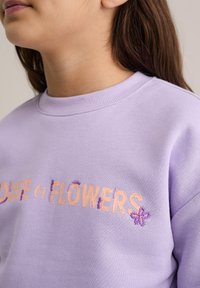 Lilaka kokvilnas sporta krekls ar izšūtu uzrakstu "HOUSE of FLOWERS" persiku krāsā ar ziedu motīviem. Rievota apkakle un mīksta tekstūra.