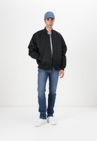 Veste bomber noire avec fermeture éclair, t-shirt gris en dessous, jean bleu, et baskets blanches. Portée avec une casquette de baseball bleu clair.