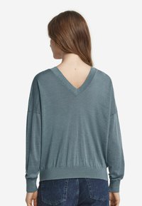 Kvinna med långt brunt hår som bär en löst sittande teal V-ringad tröja och mörkblå jeans, sedd bakifrån på en vit bakgrund.