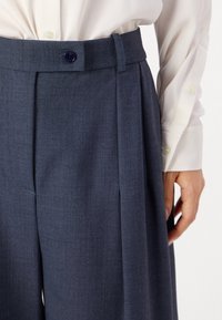 Dunkelblaue, maßgeschneiderte Hose mit weit geschnittenem Bein, Frontfalten und einem Knopfverschluss am Bund. Glatte Stofftextur und klare Linien.