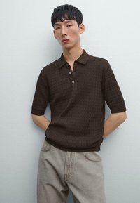 Brun strikket polo-shirt med korte ærmer, der har et struktureret mønster, en krave og en tre-knaps lukning. Kombineret med lysegrå jeans.