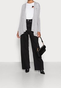 Ljusgrå, öppen cardigan över en vit t-shirt med en ficklogo, i kombination med höga svarta vida jeans och svarta klackar.