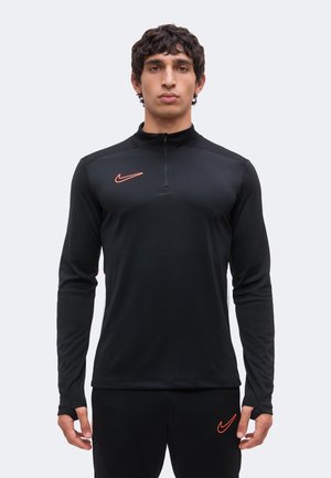 Młody mężczyzna w czarnym, długim rękawie pulowerze Nike z pomarańczowym logo, stojący przodem na jednolitym jasnym tle.
