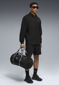 Sort lynlåssjakke, sorte shorts og sorte sneakers. Holder en sort duffelbag med hvid branding. Bærer mørke solbriller.