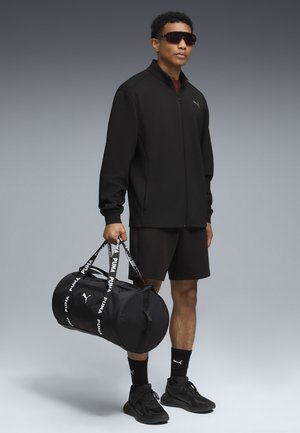 Zwarte ritssluitingjack, zwarte shorts en zwarte sneakers. Houdt een zwarte sporttas met witte branding vast. Draagt donkere zonnebrillen.