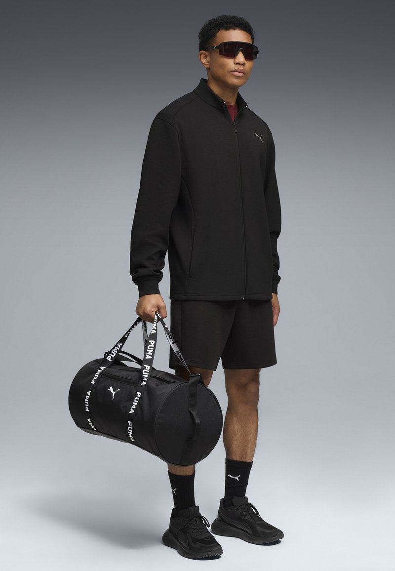 Sort lynlåssjakke, sorte shorts og sorte sneakers. Holder en sort duffelbag med hvid branding. Bærer mørke solbriller.