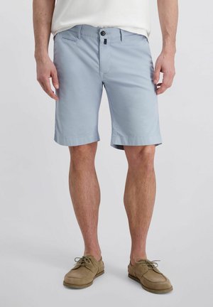 Man draagt lichtblauwe knielange shorts met knoop en rits aan de voorkant, gecombineerd met beige veterschoenen en een wit overhemd.