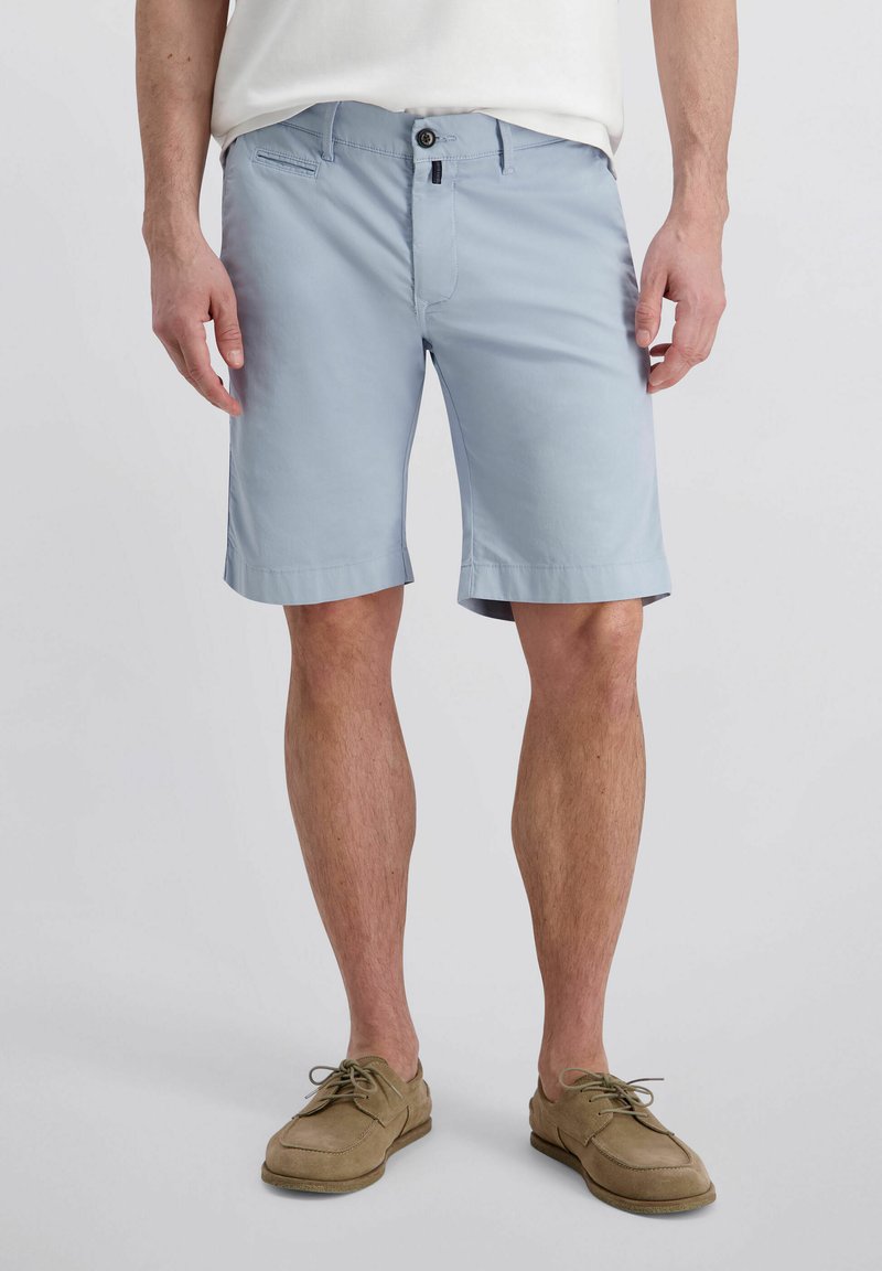 Mann trägt hellblaue knielange Shorts mit Knopf und Reißverschluss vorne, kombiniert mit hellbraunen Schnürschuhen und einem weißen Hemd.