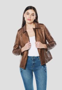 Giacca in pelle marrone con chiusura frontale con zip, dettagli trapuntati sulle spalle e colletto classico. Il modello la indossa sopra una maglia bianca e jeans blu.