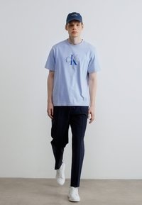 Jasnoniebieska bawełniana koszulka z nadrukowanym logo, zestawiona z ciemnymi, zwężanymi spodniami i białymi trampkami, noszona z niebieską czapką.