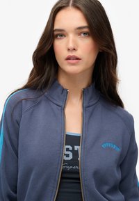 Chaqueta navy con cremallera y acentos azules, cuello acanalado y logo bordado en el pecho, combinada con una camiseta sin mangas oscura con letras grises.