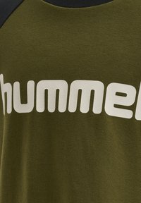 Oliwkowa zielona koszulka z czarnym wykończeniem przy dekolcie; na przodzie duży biały napis "hummel" w wyraźnej, zaokrąglonej czcionce. Gładka bawełniana faktura.