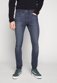 Mörkblå denimjeans med slim fit, femfickorsdesign och knappstängning i midjan. Bärs med gröna sneakers.
