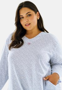 Jasnoniebieska bluzka z długim rękawem, z kwiatowym wzorem, dekoltem w serek i bocznymi kieszeniami. Z przodu znajduje się mały ozdobny akcent.