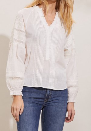 Biała bluzka z długim rękawem, z zaszewkami i koronkowymi detalami, zestawiona z niebieskimi jeansami na stojącej osobie na jednolitym tle.