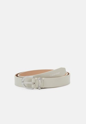 Wąski jasnobeżowy skórzany pasek z srebrną metalową klamrą i logo "Calvin Klein" blisko końca, zwinięty na białym tle.