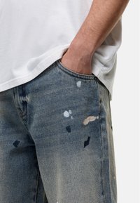 Lichtblauwe versleten denim jeans met abstracte verfvlekken en een ontspannen pasvorm. Bevat een voorzak en zichtbare stikseldetails.