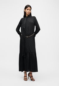 Robe noire présentant un motif texturé, des manches longues, un col à nouer et une jupe à volants, soulignée par une ceinture chaîne dorée.