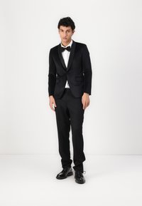 Sacou negru din tuxedo cu reveruri din satin, cămașă albă, papion negru și pantaloni negri, asortat cu pantofi negri cu șireturi. Fundal simplu.