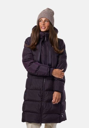 Mujer que lleva un abrigo acolchado largo de color púrpura oscuro y un gorro de punto beige, de pie con los brazos cruzados y mirando hacia un lado.
