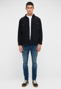 Jauns vīrietis stāv, vērsts uz priekšu, tērpies melnā korduroja žaketē virs balta krekliņa, zilos džinsos un melnās kedās ar baltām šņorēm.