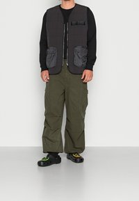 Donkergrijze gewatteerde jas met twee voorkantzakken, gedragen over een zwart shirt met lange mouwen, gecombineerd met olijfgroene cargobroeken en zwarte klompen.