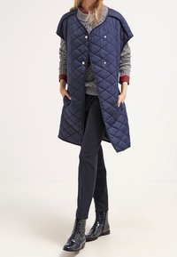 Marinblå quiltad väst med tryckknappar och vida ärmar, lagd över en grå stickad tröja och marinblå byxor, tillsammans med svarta snörstövlar.