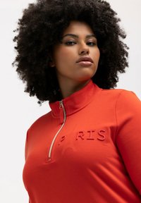 Sudadera roja con cremallera y alto cuello, que presenta el texto en relieve "PARÍS" y un sutil icono de la Torre Eiffel. Tejido suave y elástico.